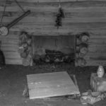 Featured image for "The History of BanjoHut.com" blog post - black and white photo depicting interior of an old shack with a fireplace, teapot, some shoes on the floor, and an old banjo hanging on the wall.