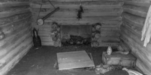 Featured image for "The History of BanjoHut.com" blog post - black and white photo depicting interior of an old shack with a fireplace, teapot, some shoes on the floor, and an old banjo hanging on the wall.