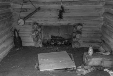 Featured image for "The History of BanjoHut.com" blog post - black and white photo depicting interior of an old shack with a fireplace, teapot, some shoes on the floor, and an old banjo hanging on the wall.